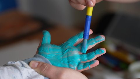 Mother or teacher preparing kid's hand to make hand prints on paper. Colourful child's hand covered with light blue paint. Slow motion 4k video