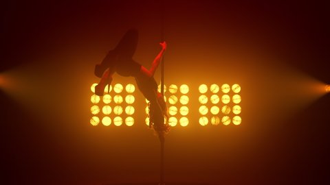 Slim woman performing poledance tricks sensually in spotlights. Sexy lady making moves upside down in night club. Young blonde dancer doing twine on pylon illuminated soft nightclub backlight.