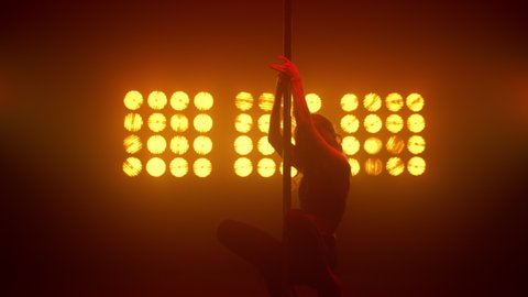 Passionate woman dancing on pole emotionally in nightclub. Hot girl performing poledance elements in strip club spotlights. Flexible blonde showing erotic dance on stage illuminated soft backlight.