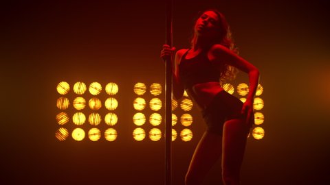 Fit girl making alluring movements performing on pole. Hot woman dancing pole dance sensually near pylon wearing sexy clothes. Young attractive lady bending body seductive in strip club backlight. 
