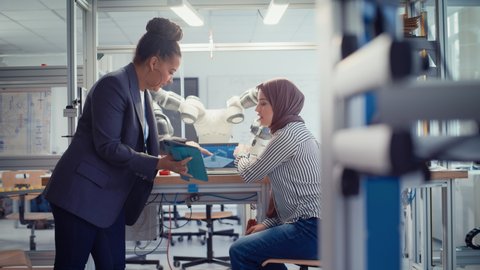 Black Senior Engineer Discussing Robotic Arm with Arabic Female Promising Computer Scientist While Manipulate it with Laptop and Tablet. Manufacturing Facility that Creates Great Products. Slow Motion