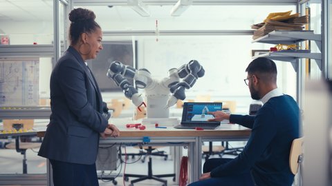 Black Chief Engineer Discussing Futuristic Robotic Arm with Young Promising Computer Scientist Man with Laptop at the Research Laboratory. Innovative Firm Startup Concept. Medium Shot