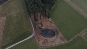A small, round pond between green fields on the edge of a coniferous forest in Germany. Aerial fly over shot