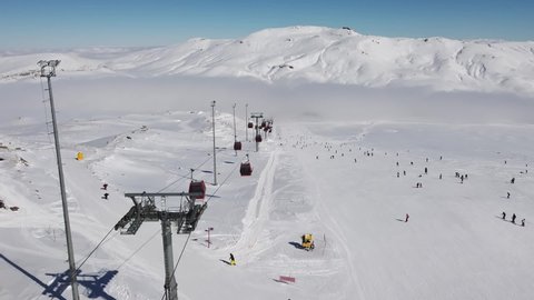 Gondola type cable car and skiers on snow covered slope of ski resort. Landscape with snowy ski piste, skiers and cloud downhill. Aerial view. Erciyes Mount, Kayseri province, Turkey