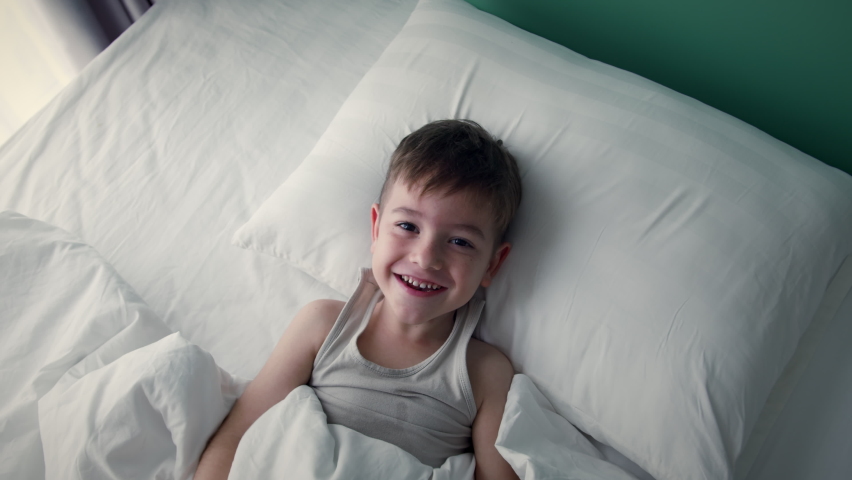 Portrait of a happy Cute funny cheerful little child boy in white pajamas wakes up in bed in the morning, looks at his parents smiling, stretches laughs and hides under the blanket again. | Shutterstock HD Video #1089452207