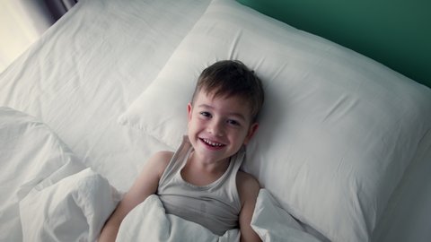 Portrait of a happy Cute funny cheerful little child boy in white pajamas wakes up in bed in the morning, looks at his parents smiling, stretches laughs and hides under the blanket again.