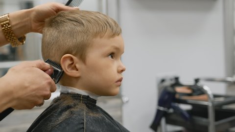 Hairdresser girl cuts the hair of blonde child with an electric trimmer machine. Process cutting hair in barber shop young girl. Men haircut and hair styling in salon. Cutting hair with trimmer.