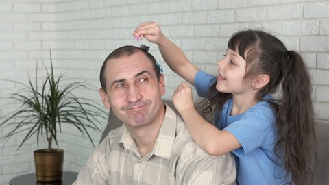 Fun with dad. A cheerful little girl rolls hairpins on her dad's head.
