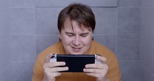 Addicted plump man plays video game console sitting in toilet at home. Focused male has fun in bathroom against grey tile wall close view.