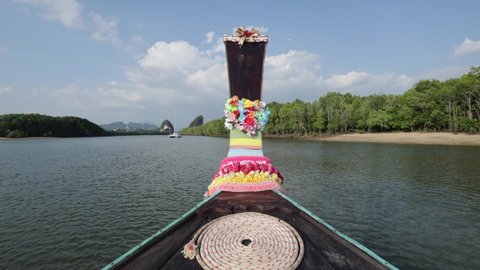 Wooden Thai longtail boat decorated with colorful flowers motoring down river during sunset with a catamaran in the distance and a white sand beach with forest on either side at Krabi Town Thailand