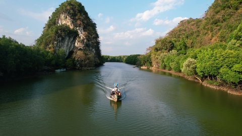 aerial drone flying tracking a thai longtail boat down river with large green limestone rocks (Khao Khanap Nam) and mangrove forests in the background on a sunset afternoon in Krabi Town Thailand