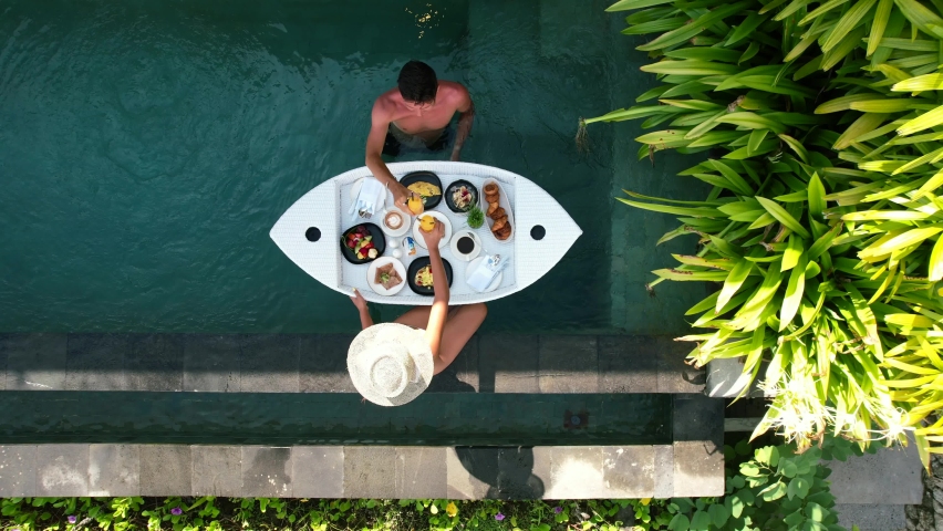 Aerial top down view of young couple with a floating breakfast poolside at a tropical luxury villa during vacation in Bali Indonesia