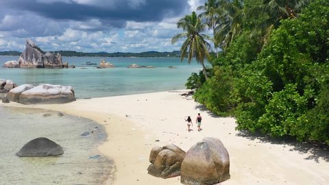 aerial of a young couple walking on a white sandy tropical beach with coconut palm trees in Belitung Indonesia on a sunny day