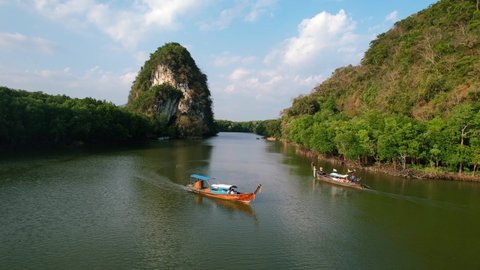 aerial of two Thai longtail boats pass each other on a green river in Krabi Town Thailand on a sunset afternoon with large limestone mountain rocks (Khao Khanap Nam) and mangroves