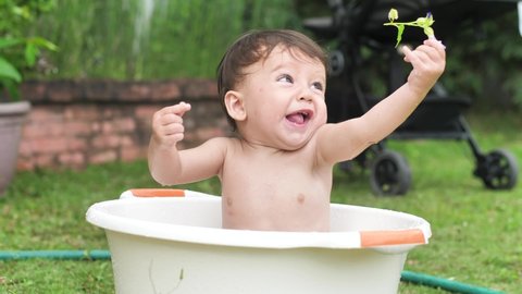 A small beautiful child sits in a round light basin, holds a flower at arm's length and looks around in surprise. Cute baby in a basin in the backyard. The baby is 8-10 months old.