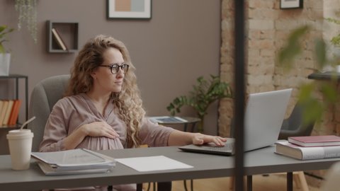 Waist-up of curly Caucasian pregnant woman in eyeglasses sitting at desk, using laptop computer in morning