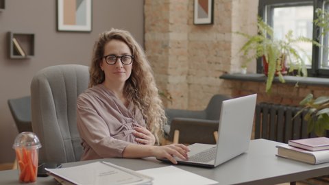 Waist up portrait of beautiful blonde pregnant woman sitting at desk in modern office at daytime, holding hand on belly and another one on laptop, looking on camera