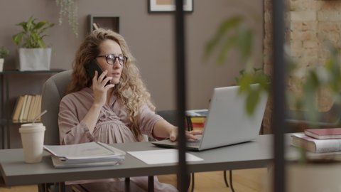 Slow motion of curly Caucasian pregnant woman in eyeglasses sitting at desk, talking on mobile phone and using laptop computer in morning