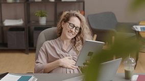 Waist-up of young long-haired Caucasian pregnant woman sitting at desk in modern office at daytime, video calling on tablet computer, smiling and waving hand - Powered by Shutterstock - Get 15% off with code: PIKWIZARD15