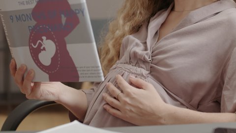 Midsection of cropped expecting woman sitting with hand on tummy in workplace at daytime, reading pregnancy literature
