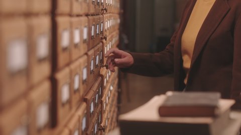 Cropped slowmo of unrecognizable female librarian opening library card index to find some information, looking through cards at wooden drawer