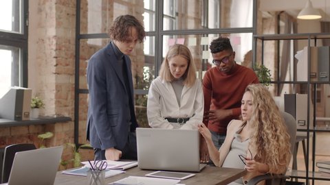 Medium of blonde expecting woman sitting at desk, Black man and Caucasian female and male colleagues standing on her side, talking and looking at portable computer