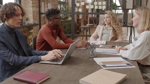 Medium of blonde pregnant woman, Black man and Caucasian female and male colleagues sitting at conference table, having business meeting and talking at daytime