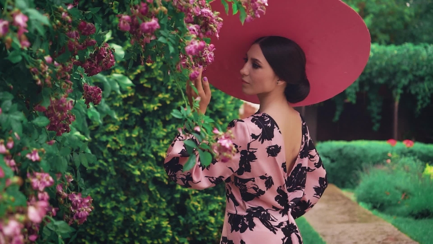 stylish pretty beauty glamour woman queen. vintage dress open sexy back. Girl princess in pink hat wide brimmed touching spring blooming green tree flowers rose bushes. Fashion model in summer nature