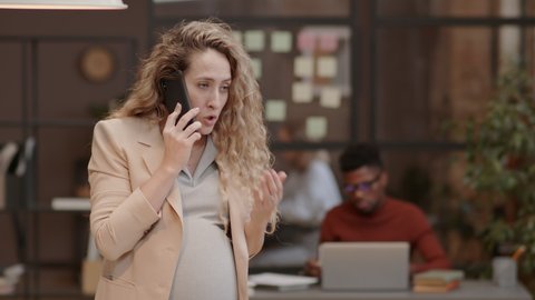 Medium of curly long-haired Caucasian mother-to-be in formalwear having stressful conversation on cellphone, standing in modern coworking space in afternoon