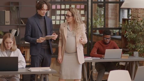 Tracking of blond-haired Caucasian mother-to-be and young man in suit walking between desks in coworking space, talking, using tablet computer