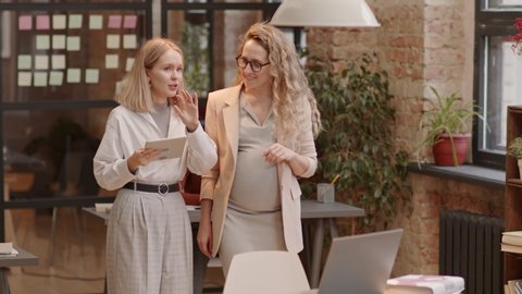 Tracking of blond-haired Caucasian mother-to-be and her young girlfriend walking between desks in coworking space, chatting, smiling