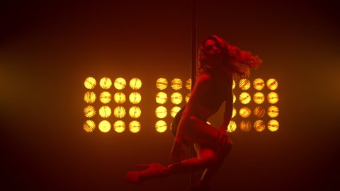 Hot woman pole dancing in front bright spotlights nightclub. Beautiful fit girl performing poledance on show stage wearing shorts. lady dancer doing erotic body moves on pylon strip club. 