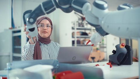 Modern Office: Portrait of Muslim Businesswoman Wearing Hijab Works on Engineering Project, Coding at Laptop and Changes Robot Hand Position. Empowered Engineer Have Startup Project
