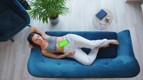 Overhead shot of relaxed woman listening to lecture or music in headphones on smartphone with green mock-up screen, lying on couch at home.