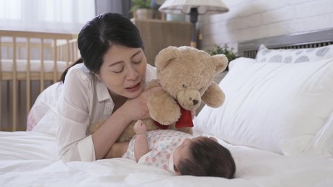 cheerful first time mother lying prone by bedside is talking to her young child with a teddy bear in hands at the bright home interior.