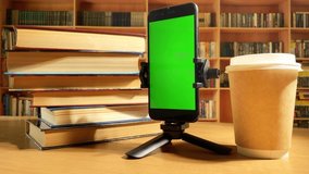 smartphone with green screen, books and paper coffee cup rotating on desk in library. Mock up display. Education concept.  Chroma key for design. 