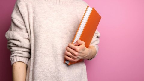 Unrecognizable woman in sweater holding book in front of pink background.