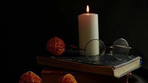 Burning candle with eyeglasses standing on old books. Big white candle burning on black background surrounded by decor. Concept of reading in dark room in illumination of flame.