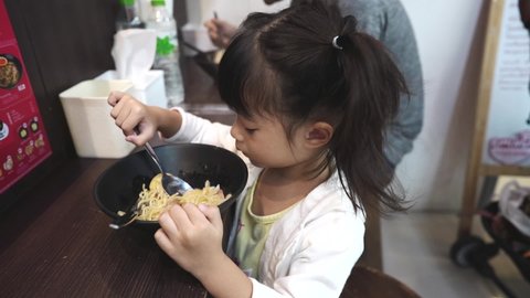 Zoom in child girl three years old self eating ramen noodle in Japanese restaurant ,she try use fork spin noodle up into her mouth. Asian child eating outside.
