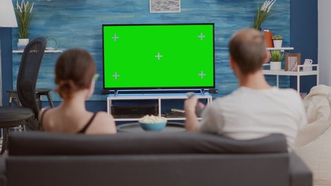 Couple sitting on couch watching television on green screen flat tv talking about favourite show and zapping in modern living room. Man and woman having popcorn enjoying movie on chroma key display.