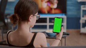 Closeup of young woman holding vertical smartphone with green screen in online conference or group video call in home living room. Static shot of girl using touchscreen mobile phone with chroma key.