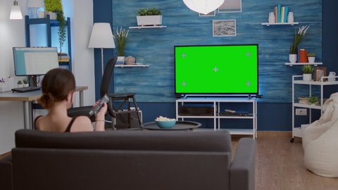 Static tripod shot of young woman zapping tv lookin for favourite show while looking at green screen in home living room. Girl relaxing using television remote changing programs on chroma key display.
