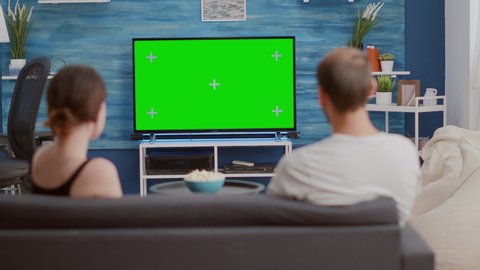 Back view of young woman asking boyfriend to change tv channel on green screen while spending time on couch. Couple enjoying television shows on chroma key mockup screen in modern living room.