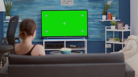 Young woman sitting on sofa looking at green screen on tv and switching channels in modern living room. Back view of girl relaxing using television remote zapping programs on chroma key display.