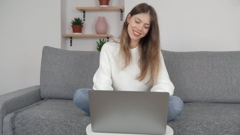 A young girl sits cross-legged on a sofa in a modern room and works on a computer. The concept of remote work or study.