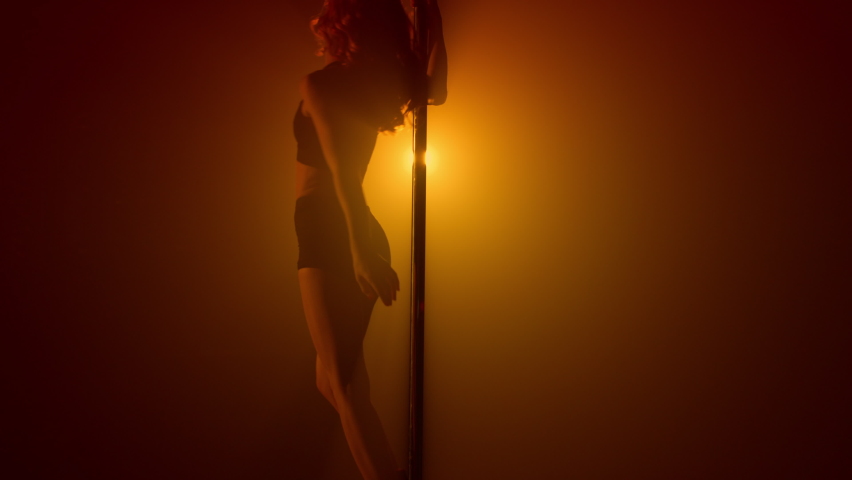 Girl dancing erotic dance at night club backlight. Fit woman practicing poledance on strip club stage. Beautiful sensual lady stripper making show on nightclub pole show illuminated soft spotlight. 