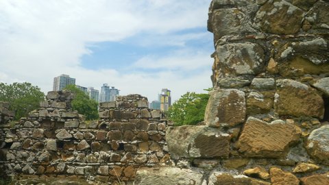 CLOSE UP: Decaying walls of ancient buildings in the green park of historic Panama Viejo obstruct view of towering skyscrapers of Panama City. High rise buildings overlook the ruins of Panama Viejo.