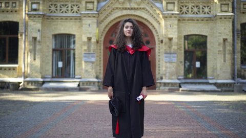 Excited satisfied beautiful woman in graduation toga jumping rejoicing looking at camera smiling. Portrait of happy Caucasian student graduating university posing at campus outdoors