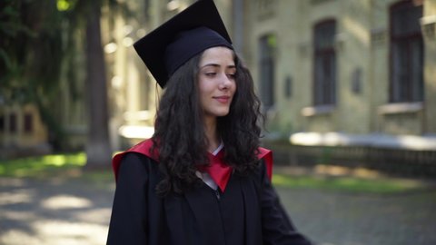 Excited intelligent beautiful woman kissing diploma standing at university campus in the morning. Portrait of smiling charming Caucasian graduate student enjoying graduation outdoors