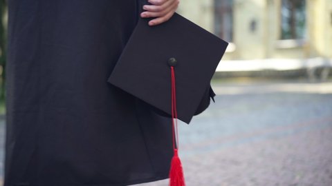 Close-up mortarboard cap in hand of unrecognizable female student walking at university campus outdoors. Young Caucasian slim woman in black graduation toga strolling at college. Tracking shot